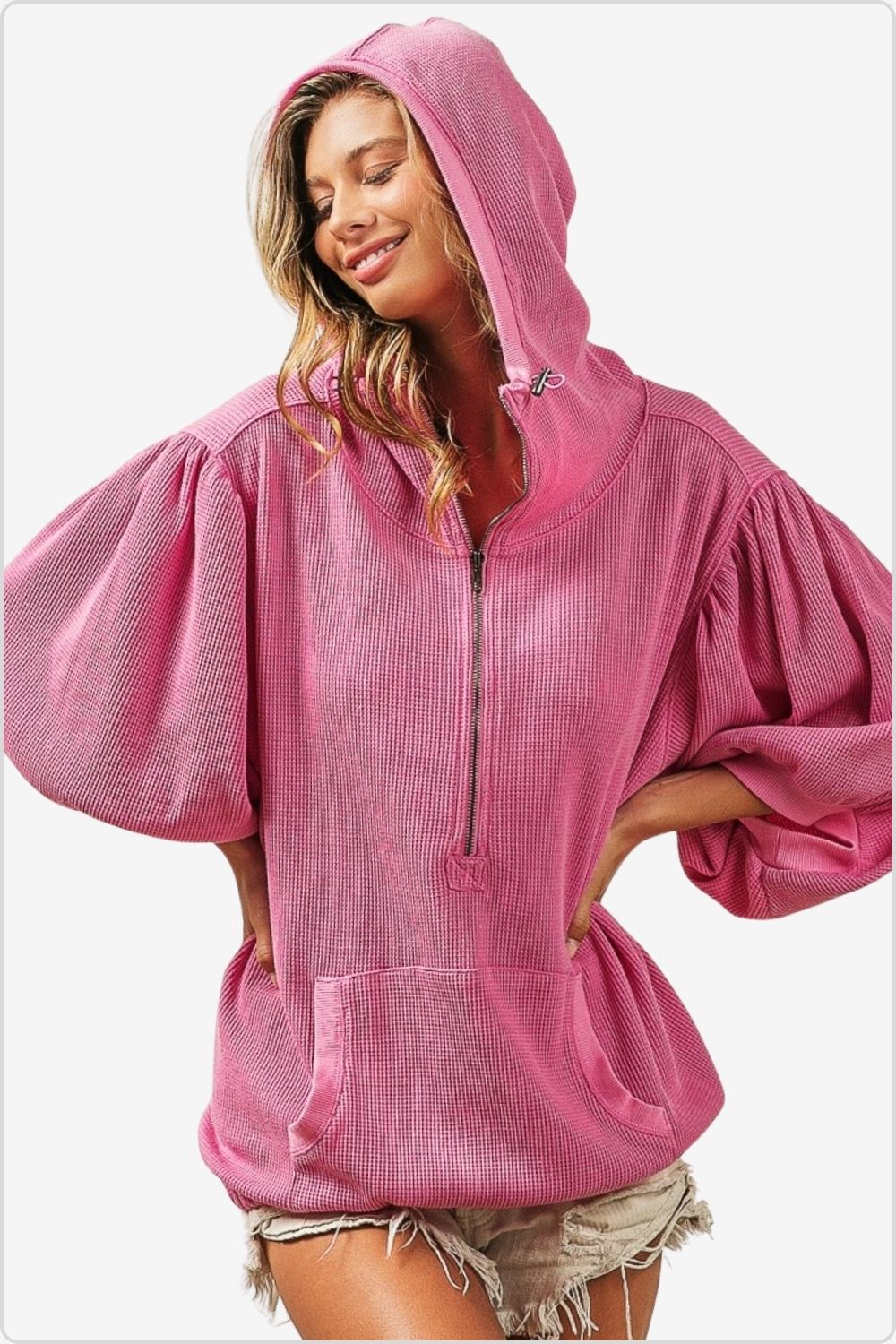 Relaxed woman in a pink waffle-knit hoodie with a half-zip front and puffed sleeves, paired with denim shorts.
