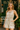 Blonde woman in plaid romper with tie neck and pockets, standing outside a green café in a springtime setting.
