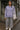 Woman wearing a light purple sweatshirt and gray pants standing on a street.