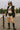 Holding skateboard in flag sweater with lace shorts, black vest, rain boots, skatepark, cloudy afternoon