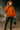 Person wearing an orange hoodie and black leather pants holding a black handbag in a dark setting.
