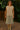 Woman wearing a beige sleeveless top and green skirt standing on a wooden deck with greenery in the background.