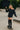 Woman in black fleece set with shorts and boat neck sweatshirt, standing on skateboard, sunny day