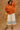 Woman wearing an orange jacket and white pants with blue patterns, standing against a brown stone wall.