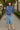 Smiling woman in blue indigo scuba sweatshirt with denim skirt and flats, walking city park path daytime.