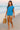 Walking barefoot on beach in blue ribbed tee with beige shorts and straw bag, soft sunset glow