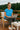Sitting on balcony in blue ribbed tee with joggers and slippers, laptop on lap, forest background, soft daylight