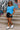 Standing on city stairs in blue ribbed tee with leather shorts and white sneakers, graffiti wall behind, daylight