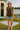 Standing near camper in brown ruffle button top with denim shorts and white slides, grassy lakeside, morning sun