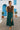 Walking on stone path in brown ruffle button top with slit teal skirt and nude sandals, white houses, midday