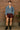 Standing in cropped denim jacket with leather shorts and lace-up boots, indoors against brick wall, soft light