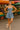 Woman wearing denim mini dress and wedge sandals, smiling near fruit and flower stands at outdoor market.