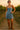 Woman wearing denim mini dress and brown suede boots, smiling on dirt path among vineyard rows at sunset.
