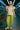 Woman in a beige top and green pants standing in a supermarket aisle with a shopping cart.