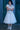 Person wearing a light-colored outfit with a skirt and sneakers, standing against a blue door.