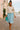Model in oatmeal linen drawstring top with pleated blue skirt and clutch, cobblestone street.