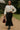 Woman walking on garden path in oatmeal linen drawstring top with black maxi skirt, velvet bag, and boots.