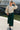Female model smiling in oatmeal linen drawstring top with green trousers, riverside bridge.
