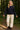 Woman in midnight eyelet cardigan with gold buttons, cream pants and boots, walking in forest path