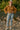 Woman in oatmeal crochet knit cardigan, blue jeans, white loafers and beige hat, standing on rocky riverbank