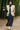 Model walking in bamboo forest in oatmeal quilted vest with charcoal wrap skirt, holding black fan, daylight