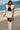 Woman posing on coastal promenade in oatmeal quilted vest with black top and white mini skirt, ocean backdrop