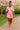 Woman in pink colorblock romper with green trim, white sandals, holding straw tote, walking by magnolias