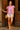 Standing in doorway in pink embroidered blouse with white shorts and round rattan bag, soft daylight