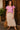 Standing by rustic door in pink embroidered blouse with tiered midi skirt and fringe bag, afternoon sun