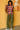 Woman wearing a colorful plaid shirt and green pants against a mustard yellow wall with abstract art.