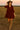 Woman walking in open field at sunset in red-brown corduroy mini dress with suede over-the-knee boots and hat