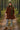Woman standing on pine forest path in red-brown corduroy mini dress with knit hat, scarf, and boots, morning light
