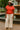 Standing by fountain in red polo sweatshirt, striped wide-leg pants and gold sandals, overcast day