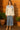 Woman in satin blouse and blue denim midi skirt with neutral heels standing on wooden terrace