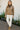 Woman wearing a brown sweater and white pants standing on a paved path with flowers in the background