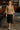 Woman walking in taupe textured knit V-neck top with black fringe skirt and heels, stylish bar interior.