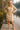 Woman in a beige sweater dress standing on a wooden dock by a lake with trees in the background