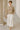 Woman standing in Wide Neck Band Drop Shoulder Sweater with wide white trousers and nude heels, marble hall daylight.