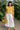 Holding fruit basket in yellow floral blouse with wide-leg pants and gold sandals, greenhouse walkway