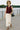 Woman walking in maroon zip front vest with cream maxi skirt and brown sandals, riverside morning.