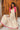 Woman wearing a white dress with pink accents sitting against a wooden wall.