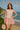 Woman in blush knit sweater and pink skirt holding a matching bag on a beach cliff