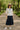 American woman in ruffled cactus blouse and navy skirt walking in a tree-lined park.