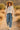 Woman in cactus blouse and light blue pants walking on a desert path in a straw hat.