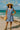 Ruffle dress styled with lace-up green sandals, straw hat, and round raffia crossbody by the ocean