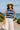 Woman in knit crop tank and wide-leg white pants with hat on a coastal path