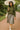 Smiling woman walking in army green mock neck sweater with pleated skirt and sneakers, garden path daytime