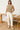 Woman wearing a beige sleeveless sweater and white pants standing in a casual setting with wooden furniture.