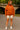 Person wearing an orange hoodie with a zipper, white shorts, and white sneakers in a park setting.