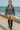 Model walking by ocean in charcoal ribbed half zip sweater with silver shorts and thigh high boots, cloudy shore.